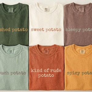 May include: Six crew neck t-shirts in olive green, cream, mauve, sage green, terracotta, and mustard yellow. Each shirt features text printed on the chest: mashed potato, sweet potato, sleepy potato, couch potato, kind of rude potato, and spicy potato.