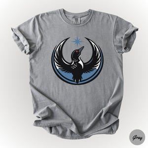 K&ouml;nnte beinhalten: Graues T-Shirt mit einer Grafik eines schwarzen Vogels mit ausgebreiteten Fl&uuml;geln, einem roten Auge und einem blauen Stern dar&uuml;ber. Der Vogel befindet sich in einem blauen und schwarzen Kreis. Das Shirt besteht aus einem weichen Material.