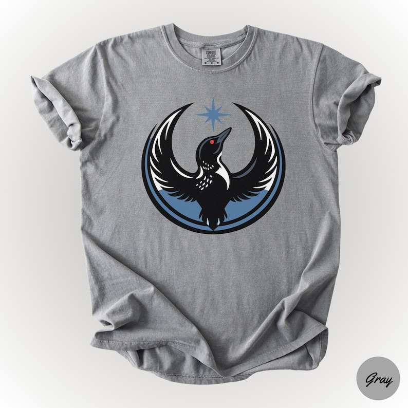 K&ouml;nnte beinhalten: Graues T-Shirt mit einer Grafik eines schwarzen Vogels mit ausgebreiteten Fl&uuml;geln, einem roten Auge und einem blauen Stern dar&uuml;ber. Der Vogel befindet sich in einem blauen und schwarzen Kreis. Das Shirt besteht aus einem weichen Material.