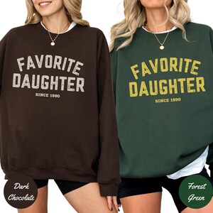 K&ouml;nnte beinhalten: Zwei Sweatshirts, eines in Dunkelschokolade und eines in Waldgr&uuml;n, jeweils mit dem Text "FAVORITE DAUGHTER SINCE 1990" in einer Used-Schriftart. Das dunkelbraune Sweatshirt hat wei&szlig;en Text, das gr&uuml;ne Sweatshirt gelben Text.