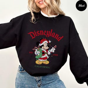 K&ouml;nnte beinhalten: Schwarzes Disneyland-Sweatshirt mit einem festlichen Mickey Mouse-Design. Mickey tr&auml;gt einen Weihnachtsmann-Anzug und h&auml;lt Zuckerstangen und einen Sack. Das Wort "Disneyland" steht in roter Schrift &uuml;ber der Grafik, darunter "Est. 1955" und "California".