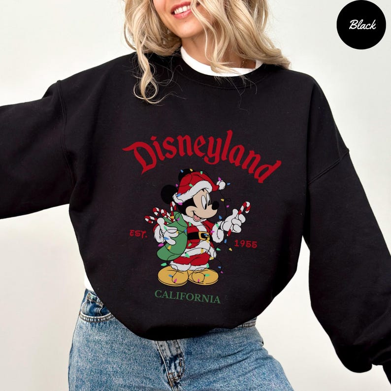 K&ouml;nnte beinhalten: Schwarzes Disneyland-Sweatshirt mit einem festlichen Mickey Mouse-Design. Mickey tr&auml;gt einen Weihnachtsmann-Anzug und h&auml;lt Zuckerstangen und einen Sack. Das Wort "Disneyland" steht in roter Schrift &uuml;ber der Grafik, darunter "Est. 1955" und "California".