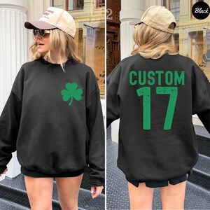 K&ouml;nnte beinhalten: Schwarzes Sweatshirt mit einem gr&uuml;nen Kleeblatt auf der Vorderseite. Auf der R&uuml;ckseite des Sweatshirts steht das Wort "CUSTOM" &uuml;ber der Zahl "17" in Gr&uuml;n. Das Sweatshirt hat einen l&auml;ssigen Stil.