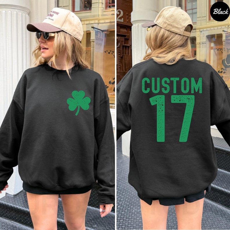K&ouml;nnte beinhalten: Schwarzes Sweatshirt mit einem gr&uuml;nen Kleeblatt auf der Vorderseite. Auf der R&uuml;ckseite des Sweatshirts steht das Wort "CUSTOM" &uuml;ber der Zahl "17" in Gr&uuml;n. Das Sweatshirt hat einen l&auml;ssigen Stil.