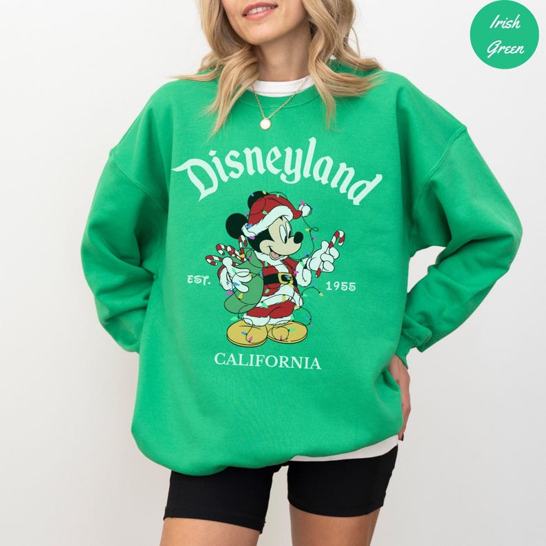 K&ouml;nnte beinhalten: Gr&uuml;nes Disneyland-Sweatshirt mit einer Grafik von Mickey Mouse in einem Weihnachtsmannhut und -anzug, der Zuckerstangen h&auml;lt. Auf dem Sweatshirt stehen die W&ouml;rter "Disneyland" und "California", sowie das Jahr "1955" und das Wort "EST."
