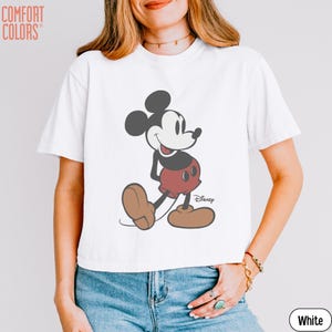 May include: White t-shirt featuring a classic Mickey Mouse graphic. The cartoon character is depicted in black, red, and brown, with the Disney logo. The shirt is a Comfort Colors brand.