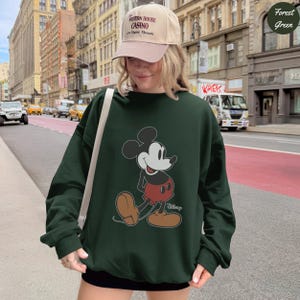 Puede incluir: Sudadera verde bosque con un gr&aacute;fico de Mickey Mouse. El personaje de dibujos animados se representa caminando, con pantalones cortos rojos y zapatos marrones. La persona lleva una gorra beige con texto. El fondo muestra una calle de la ciudad.