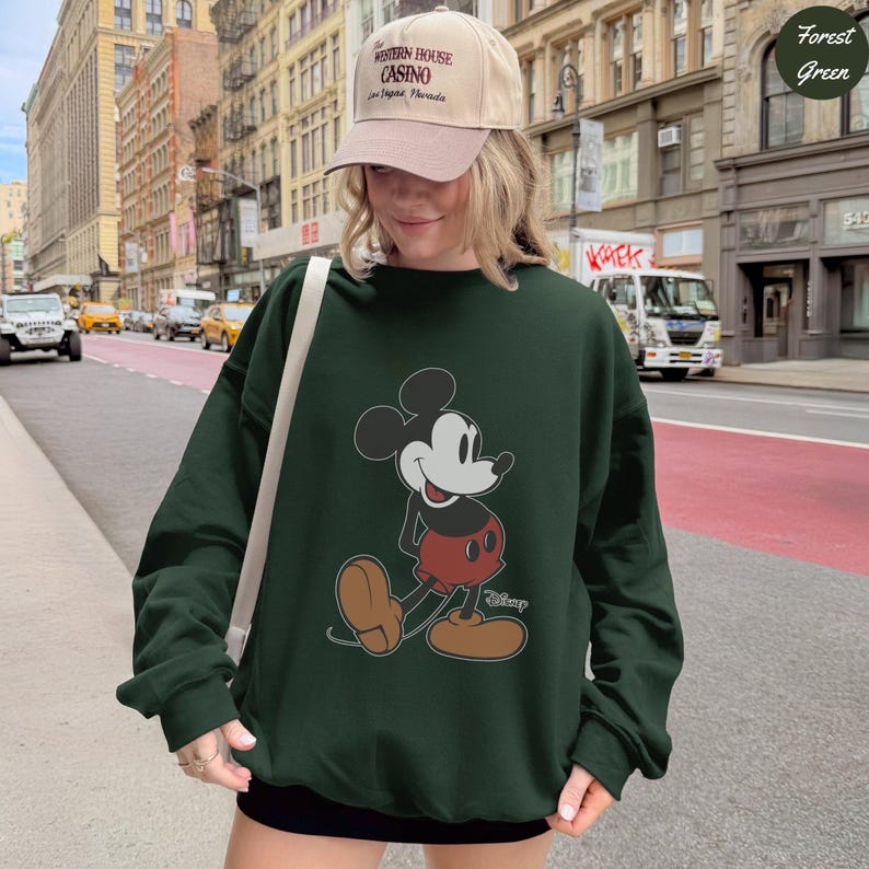 Puede incluir: Sudadera verde bosque con un gr&aacute;fico de Mickey Mouse. El personaje de dibujos animados se representa caminando, con pantalones cortos rojos y zapatos marrones. La persona lleva una gorra beige con texto. El fondo muestra una calle de la ciudad.