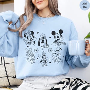 May include: Light blue sweatshirt featuring black and white illustrations of Disney characters, including Mickey Mouse, Minnie Mouse, Pluto, Daisy Duck, Goofy, and Donald Duck. The text "Light Blue" is in a circle in the upper right corner.