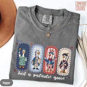 May include: A grey Comfort Colors t-shirt with a patriotic goose design. The design shows four cartoon geese dressed as the Statue of Liberty, a Founding Father, Uncle Sam, and a Revolutionary War soldier. The text "Just a patriotic goose" is printed below the geese.