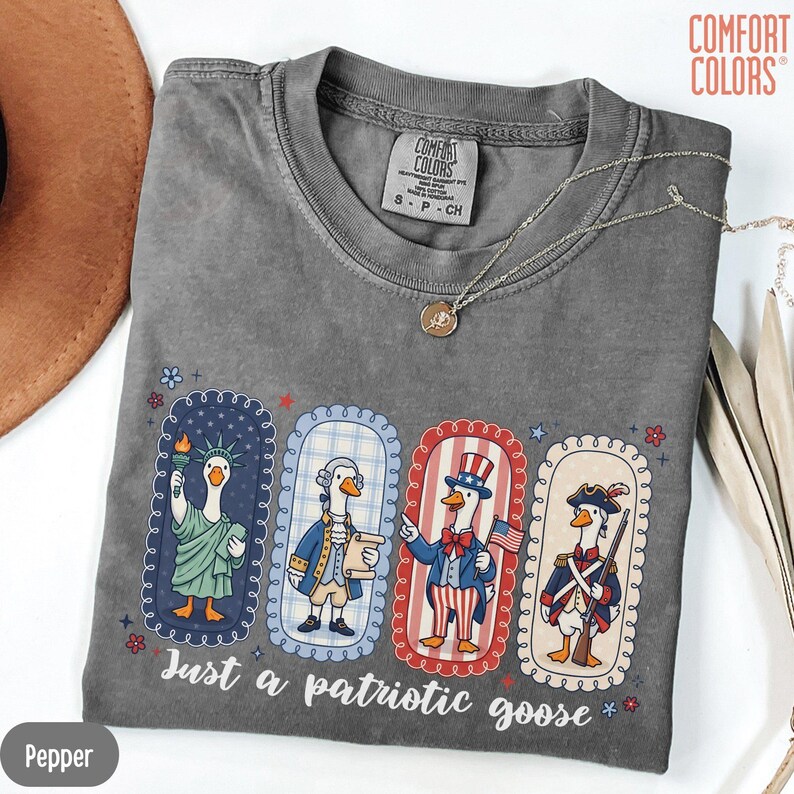May include: A grey Comfort Colors t-shirt with a patriotic goose design. The design shows four cartoon geese dressed as the Statue of Liberty, a Founding Father, Uncle Sam, and a Revolutionary War soldier. The text "Just a patriotic goose" is printed below the geese.