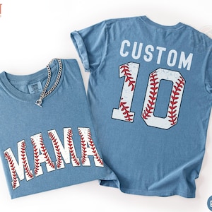 May include: Two blue jean-colored t-shirts. One shirt has the word "NANA" in white letters with red baseball stitching. The other shirt has the word "CUSTOM" above the number "10" in white with red baseball stitching. A silver chain necklace is on the first shirt.