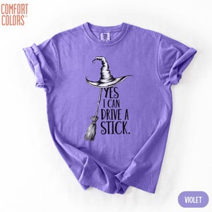 Puede incluir: Una camiseta violeta de manga corta con un gr&aacute;fico negro de un sombrero de bruja sobre una escoba. El texto "YES I CAN DRIVE A STICK." est&aacute; impreso en letras negras junto a la escoba. La camiseta tiene cuello redondo y mangas remangadas.