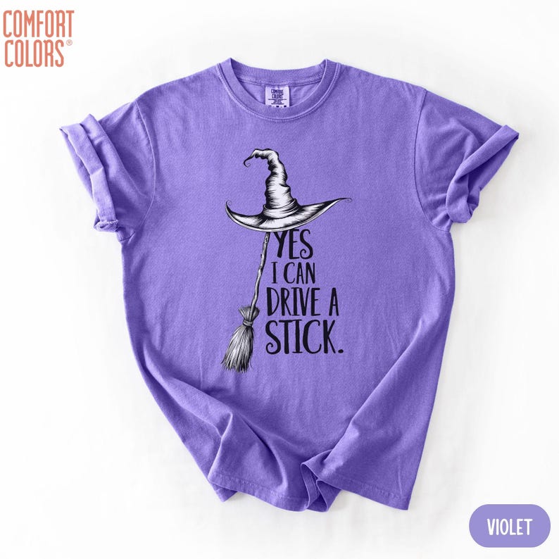 Puede incluir: Una camiseta violeta de manga corta con un gr&aacute;fico negro de un sombrero de bruja sobre una escoba. El texto "YES I CAN DRIVE A STICK." est&aacute; impreso en letras negras junto a la escoba. La camiseta tiene cuello redondo y mangas remangadas.