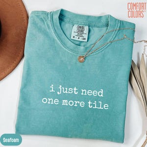 Pode incluir: Camiseta Comfort Colors verde-água com o texto "i just need one more tile" em branco. Um colar de ouro com um pingente repousa sobre a camisa. A camisa está dobrada e exibida com um chapéu marrom.