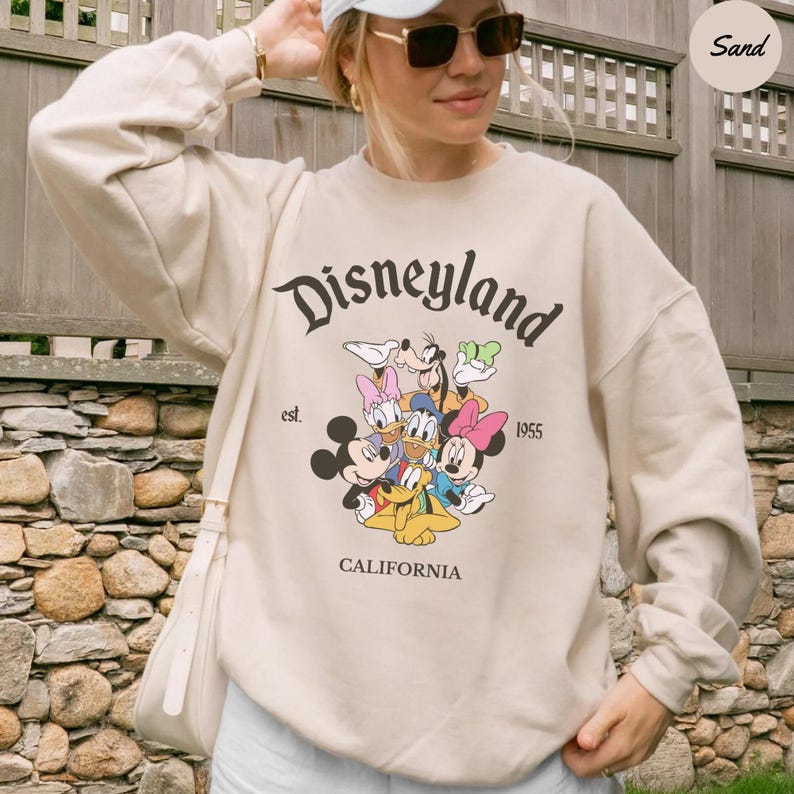 K&ouml;nnte beinhalten: Beiges Disneyland-Sweatshirt mit Mickey Mouse, Minnie Mouse, Donald Duck, Daisy Duck, Goofy und Pluto. Das Sweatshirt zeigt das Disneyland-Logo, "est. 1955" und "CALIFORNIA" auf der Vorderseite.
