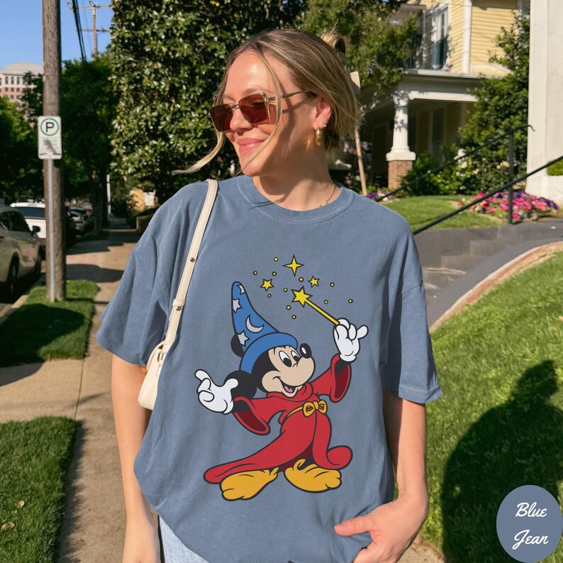 May include: Blue t-shirt featuring a cartoon image of Mickey Mouse dressed as a wizard, holding a magic wand with stars. The shirt is a faded blue color and has a relaxed fit. The text "Blue Jean" is in the bottom right corner.