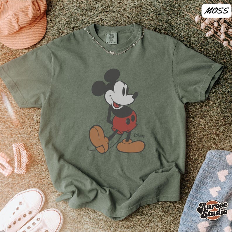 May include: Moss green t-shirt featuring a classic Mickey Mouse graphic. The cartoon character is depicted in his signature red shorts, large yellow shoes, and white gloves. The shirt has a crew neck and short sleeves. A small Disney logo is visible.
