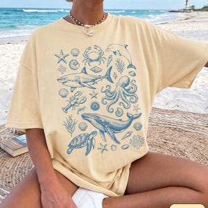 Pode incluir: Uma t-shirt de manga curta na cor manteiga com uma estampa gr&aacute;fica azul de v&aacute;rias criaturas marinhas, incluindo um tubar&atilde;o, baleia, polvo, tartaruga marinha, caranguejo, golfinho, lagosta, estrelas-do-mar e conchas dispostas em um padr&atilde;o de colagem.