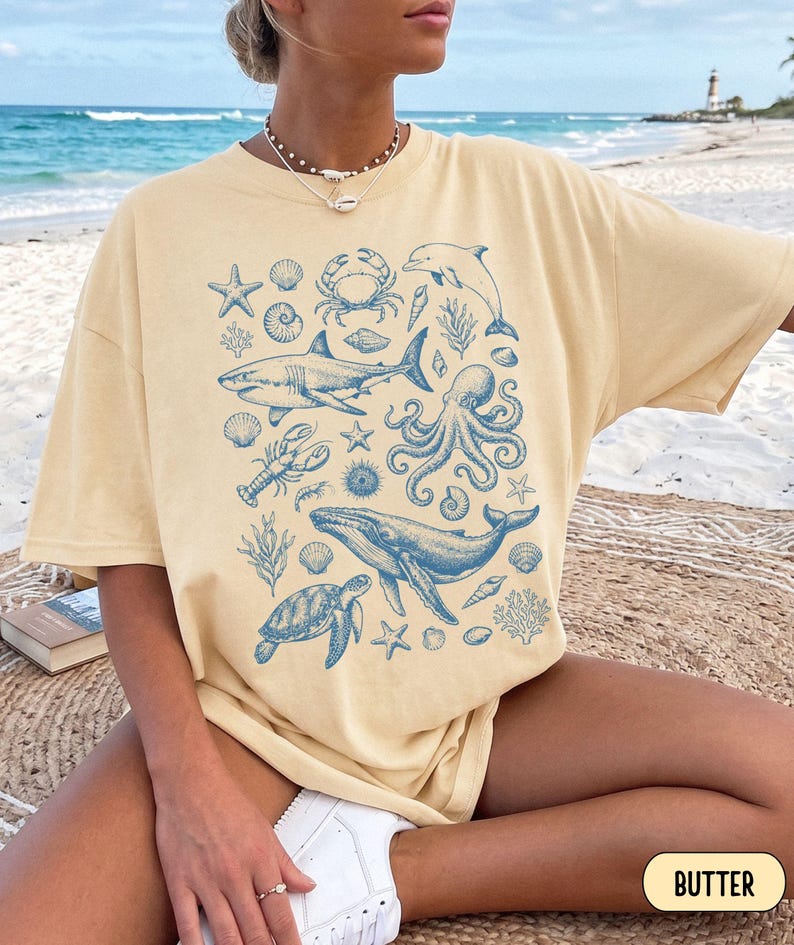 Pode incluir: Uma t-shirt de manga curta na cor manteiga com uma estampa gr&aacute;fica azul de v&aacute;rias criaturas marinhas, incluindo um tubar&atilde;o, baleia, polvo, tartaruga marinha, caranguejo, golfinho, lagosta, estrelas-do-mar e conchas dispostas em um padr&atilde;o de colagem.