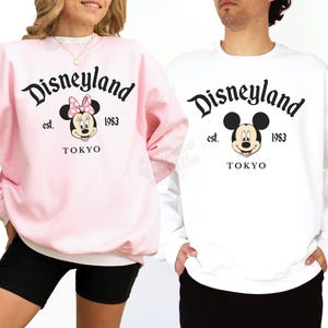 May include: Two sweatshirts, one light pink and one white, featuring the Disneyland logo with Mickey and Minnie Mouse. The text includes "est. 1983" and "TOKYO". The Minnie Mouse design has a pink bow.