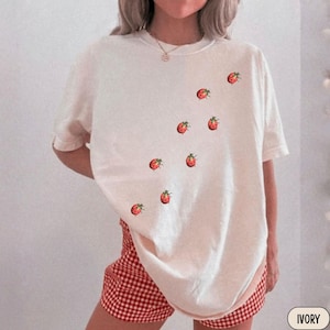 May include: An ivory short-sleeved t-shirt featuring a scattered pattern of small red and black ladybug illustrations across the front. The shirt has a crew neckline and a loose, relaxed fit, paired with red and white checkered shorts.