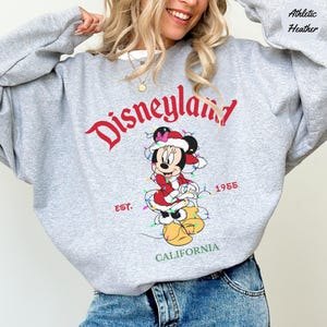 K&ouml;nnte beinhalten: Graues Disneyland Sweatshirt mit Minnie Mouse im Weihnachtskost&uuml;m und Lichterkette. Der Schriftzug "Disneyland" ist rot, darunter steht "EST. 1955 CALIFORNIA".