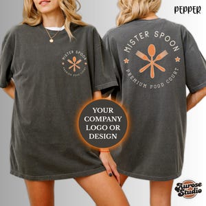 May include: Pepper-colored t-shirts with the text "Mister Spoon Premium Food Court" and a logo featuring a spoon, fork, and knife. The shirts have a vintage look and are worn by a person. The front of the shirt has a smaller version of the logo.