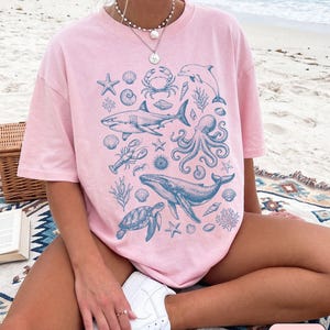 Pode incluir: Uma t-shirt de manga curta cor-de-rosa claro com uma impress&atilde;o gr&aacute;fica azul de v&aacute;rias criaturas marinhas, incluindo um tubar&atilde;o, uma baleia, um polvo, uma tartaruga marinha, um caranguejo, uma lagosta, um golfinho, estrelas-do-mar e conchas.