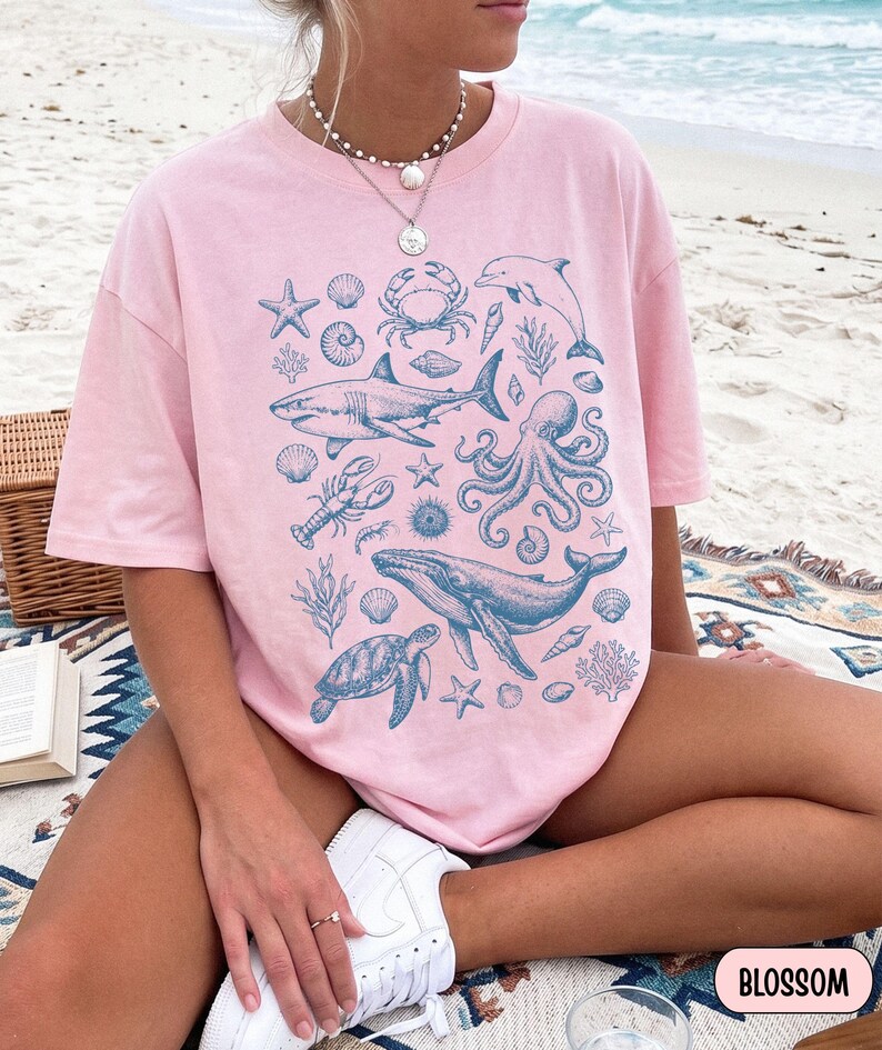 Pode incluir: Uma t-shirt de manga curta cor-de-rosa claro com uma impress&atilde;o gr&aacute;fica azul de v&aacute;rias criaturas marinhas, incluindo um tubar&atilde;o, uma baleia, um polvo, uma tartaruga marinha, um caranguejo, uma lagosta, um golfinho, estrelas-do-mar e conchas.