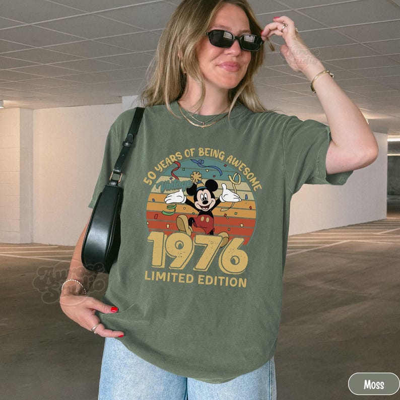 Puede incluir: Camiseta verde oliva con un dise&ntilde;o de Mickey Mouse y el texto "50 YEARS OF BEING AWESOME 1976 LIMITED EDITION". El dise&ntilde;o est&aacute; sobre un fondo retro de puesta de sol.
