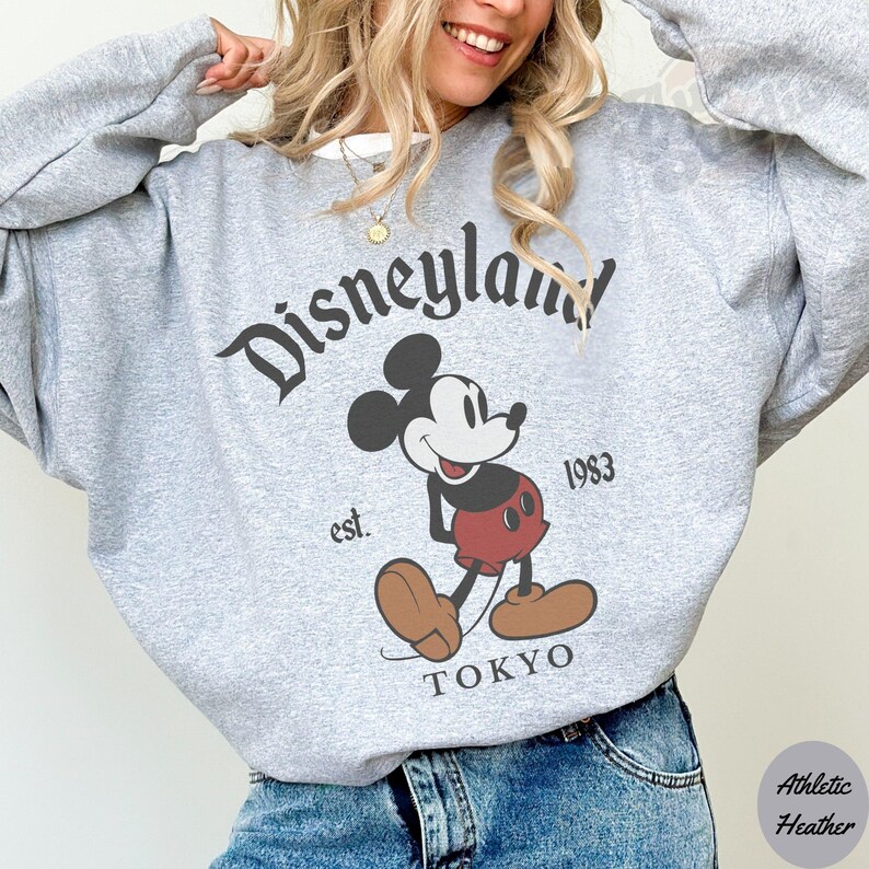 Puede incluir: Una sudadera gris con la palabra "Disneyland" en una fuente vintage y un gr&aacute;fico de Mickey Mouse. La sudadera tambi&eacute;n incluye el texto "est. 1983 TOKYO". La tela es athletic heather.