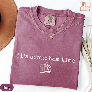 Puede incluir: Camiseta color bayas con la frase "it's about bam time" en blanco. La camiseta presenta un gráfico de dos fichas de Mahjong. Un collar de oro con un colgante descansa sobre la camiseta.