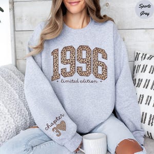 Op de afbeelding: Een lichtgrijs sweatshirt met het jaar "1996" in een luipaardprint ontwerp. De tekst "limited edition" staat onder het jaar. De linkermouw heeft de tekst "chapter 39" met een luipaardprint hart.