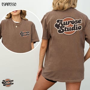 May include: Espresso-colored t-shirt with the text "Aurose Studio" on the front and back. The logo is in a retro style with brown and cream colors. The shirt is a short-sleeved, crew neck style.