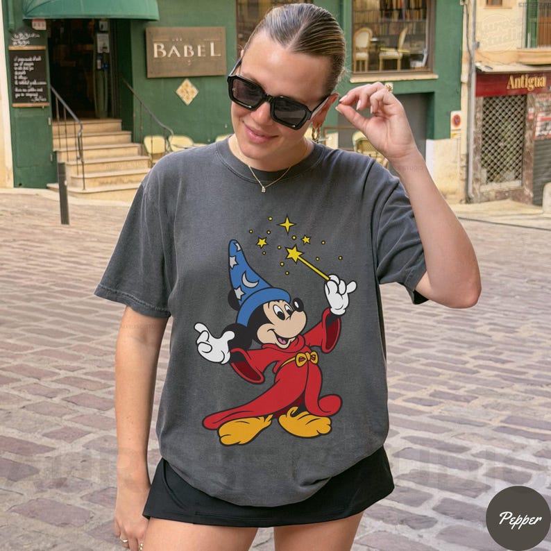 May include: Dark gray t-shirt featuring a graphic of Mickey Mouse dressed as a sorcerer, holding a wand with stars. The cartoon character is wearing a blue hat, red robe, and yellow shoes. The shirt is a relaxed fit.