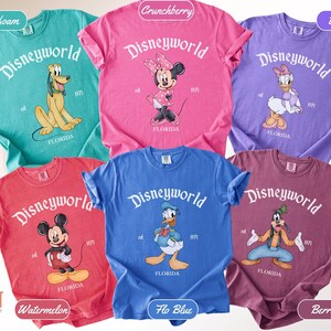 May include: A collection of Comfort Colors t-shirts in various colors, each featuring a Disney World graphic. The shirts display different Disney characters, including Mickey Mouse, Minnie Mouse, Donald Duck, Pluto, Daisy Duck, and Goofy. The text "Disneyworld" and "Florida" are printed on each shirt.