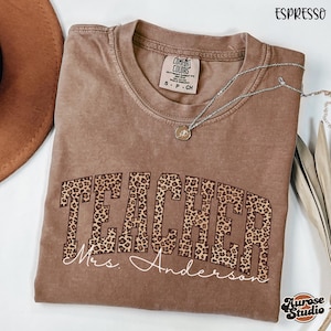 Könnte beinhalten: Espressofarbenes T-Shirt mit dem Wort "TEACHER" in einem Leopardenmuster. Der Name "Mrs. Anderson" ist unter dem Wort "TEACHER" in kursiver Schrift gedruckt. Eine silberne Halskette mit Anhänger befindet sich auf dem Shirt.