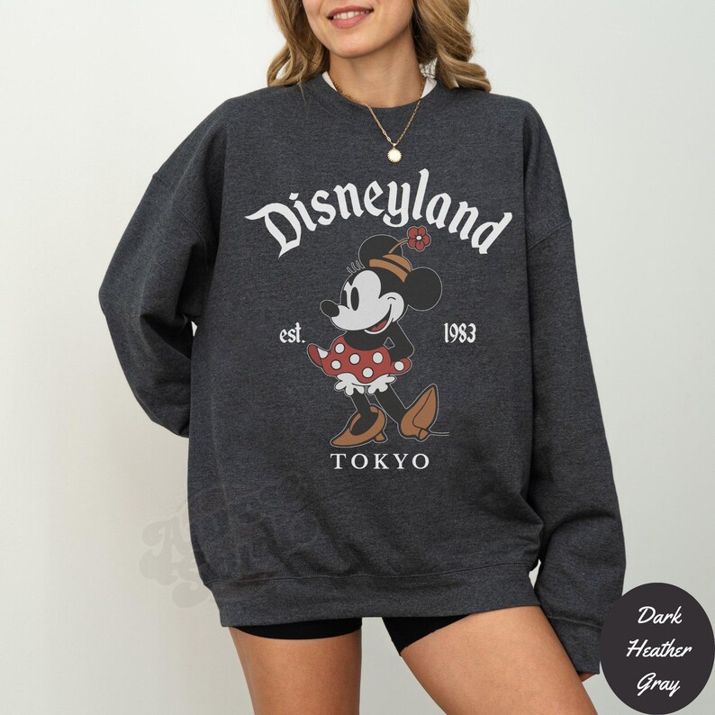 Puede incluir: Sudadera gris oscuro con un gr&aacute;fico de Minnie Mouse. El dise&ntilde;o incluye el texto "Disneyland Tokyo est. 1983". Minnie Mouse se representa con un vestido de lunares rojos y zapatos marrones. La sudadera es de estilo casual.