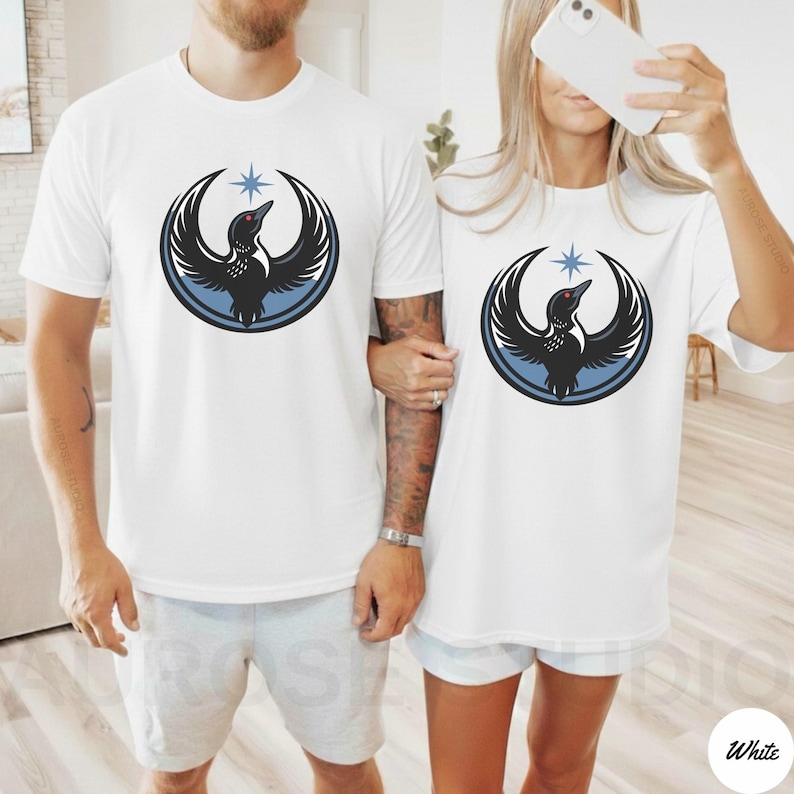 K&ouml;nnte beinhalten: Wei&szlig;e T-Shirts mit einem Grafikdesign eines schwarzen Vogels mit ausgebreiteten Fl&uuml;geln, einem blauen Kreis und einem Stern. Das Vogeldesign befindet sich mittig auf der Vorderseite der Shirts. Die Shirts werden von zwei Personen getragen.