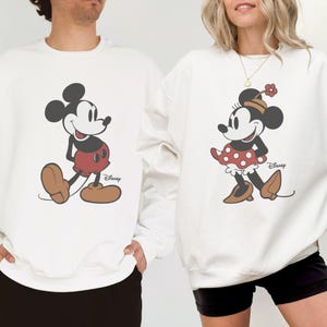 Puede incluir: Sudaderas blancas con Mickey y Minnie Mouse. Mickey lleva pantalones cortos rojos y zapatos marrones. Minnie lleva una falda de lunares rojos, zapatos marrones y una flor en la cabeza. El logotipo de Disney est&aacute; impreso en la parte inferior derecha.