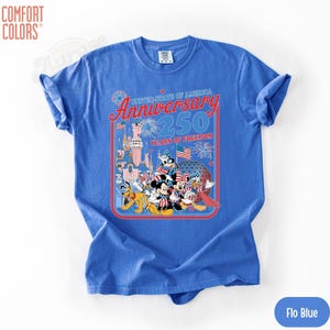 May include: A blue Comfort Colors t-shirt featuring a graphic celebrating the United States of America's 250th Anniversary. The design includes cartoon characters, fireworks, flags, and a castle. The shirt is labeled "Flo Blue".