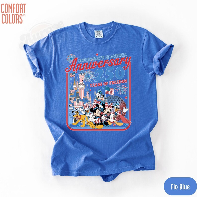 May include: A blue Comfort Colors t-shirt featuring a graphic celebrating the United States of America's 250th Anniversary. The design includes cartoon characters, fireworks, flags, and a castle. The shirt is labeled "Flo Blue".