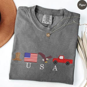 May include: Gray Comfort Colors t-shirt with embroidered USA-themed designs: a cowboy boot, American flag, eagle, and red truck. A silver necklace with a pendant is draped across the shirt.