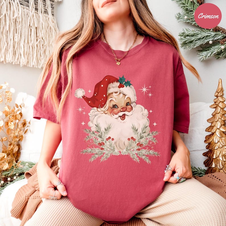 May include: Crimson-colored t-shirt featuring a vintage Santa Claus graphic. Santa is depicted with a red hat, white beard, and a smiling face, surrounded by sprigs of greenery and berries. The design includes star accents, creating a festive holiday theme.