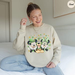 May include: Beige sweatshirt featuring Mickey Mouse, Pluto, and Minnie Mouse in St. Patrick's Day attire. Mickey wears a green hat, Minnie a green bow, and Pluto a green collar. Gold coins and shamrocks decorate the design. The word "Lucky" is on the design.