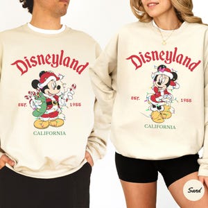 K&ouml;nnte beinhalten: Beige Sweatshirts mit dem Disneyland-Logo in roter Schrift. Micky Maus in einem Weihnachtsmann-Outfit ist unter dem Logo abgebildet, mit dem Text "EST. 1955 CALIFORNIA". Die Sweatshirts sind ein festliches Weihnachtsdesign.
