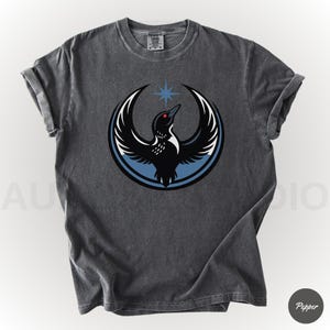 K&ouml;nnte beinhalten: Dunkelgraues T-Shirt mit einem kreisf&ouml;rmigen Design, das einen schwarzen Vogel mit ausgebreiteten Fl&uuml;geln, einem roten Auge und einem blauen Stern zeigt. Der Vogel ist vor einem blau-wei&szlig;en Hintergrund platziert. Die &Auml;rmel sind hochgekrempelt.