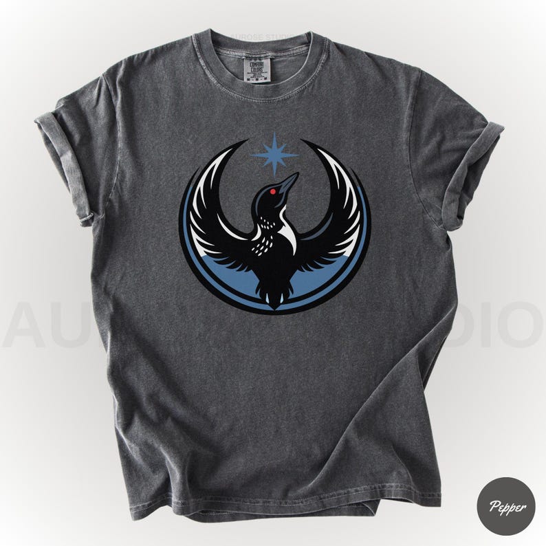 K&ouml;nnte beinhalten: Dunkelgraues T-Shirt mit einem kreisf&ouml;rmigen Design, das einen schwarzen Vogel mit ausgebreiteten Fl&uuml;geln, einem roten Auge und einem blauen Stern zeigt. Der Vogel ist vor einem blau-wei&szlig;en Hintergrund platziert. Die &Auml;rmel sind hochgekrempelt.