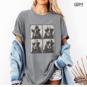 Op de afbeelding: Grijze t-shirt met een vierpanelen grafische afbeelding van kippen met zonnebrillen. Het shirt heeft een relaxte pasvorm en opgerolde mouwen. Het woord "Gray" staat rechtsboven gedrukt. De merknaam "Aurose Studio" staat rechtsonder gedrukt.